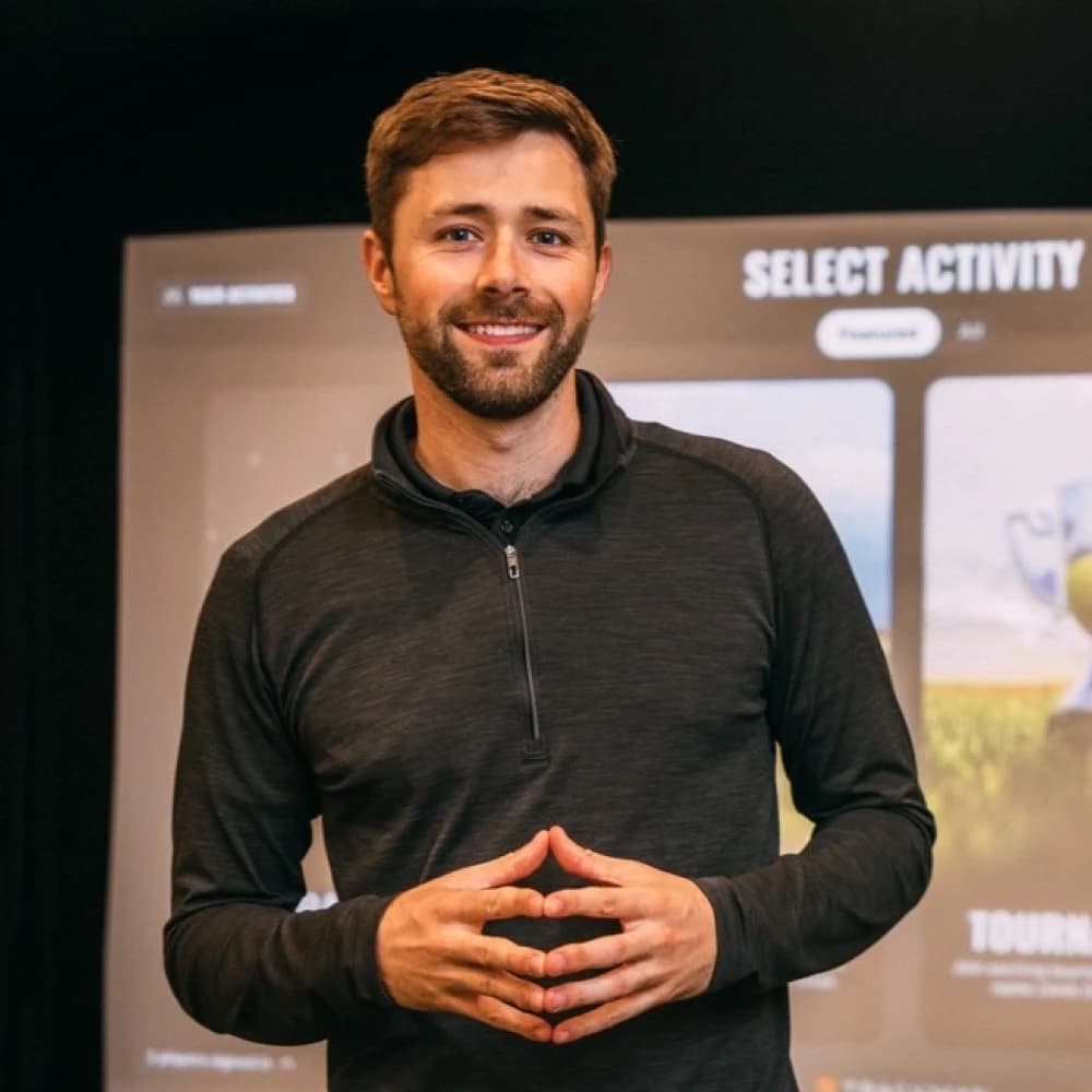 Man in a dark shirt stands in front of a screen displaying "Select Activity," smiling with hands together.