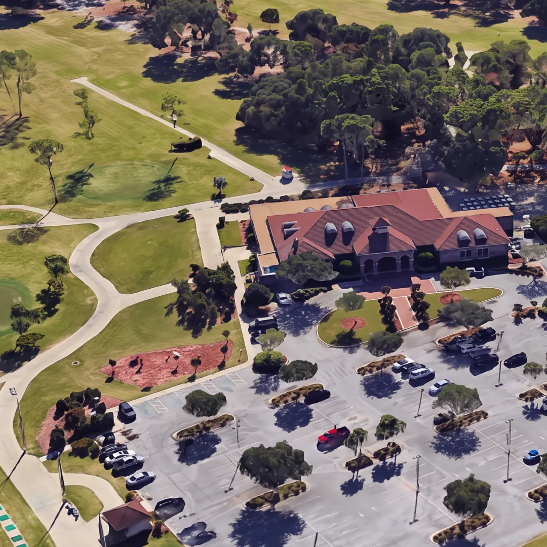 Aerial view of a golf course with a clubhouse, surrounded by parking lots and green fairways, bordered by trees and pathways.