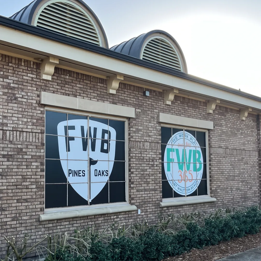 A brick building with two large signs on the windows, labeled "FWB Pines Oaks" and "FWB 365," with plants below the windows.