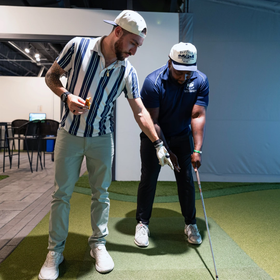 Two people on a golf simulator, one in a striped shirt helping the other with a golf club grip. Both wearing caps, standing on an artificial turf.