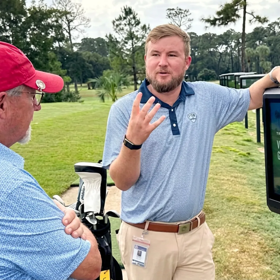 Two men are talking near a golf course; one gestures while the other listens. Golf clubs and trees are visible in the background.