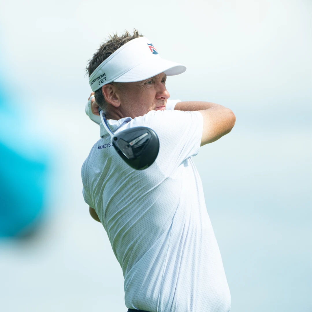 Golfer in a white shirt and visor swings his club on a bright day, focused on the shot.