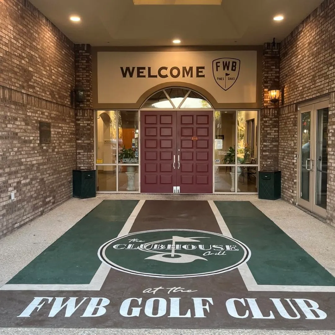 Entrance to the FWB Golf Club with large red double doors, a welcome sign above, and clubhouse grill logo on the walkway.