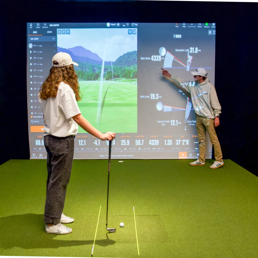 Two people in a golf simulator room with data displayed on a screen, one holding a club and the other pointing at the screen.