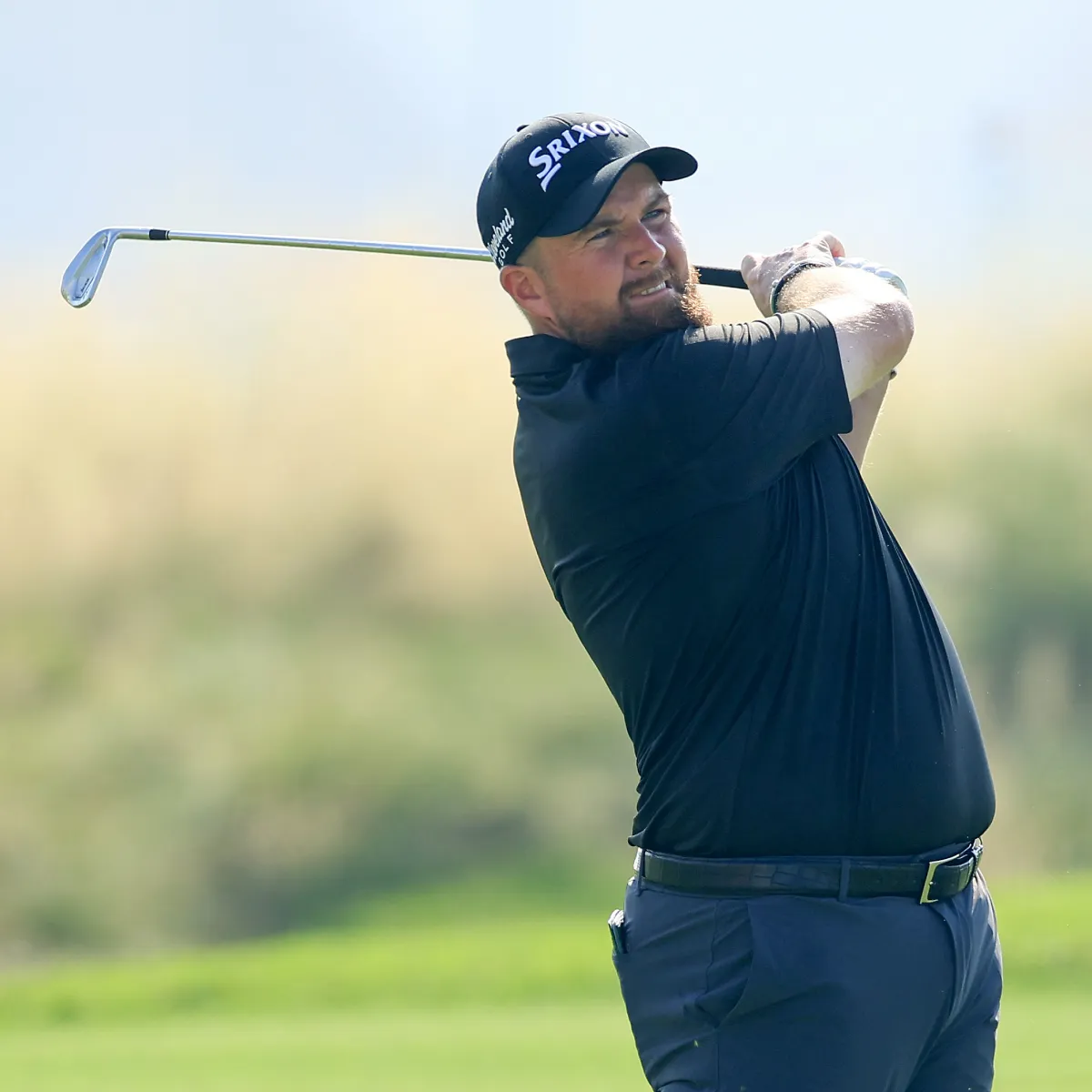 A golfer in a black shirt and cap swings a club on a sunny day, with a blurred grassy background.