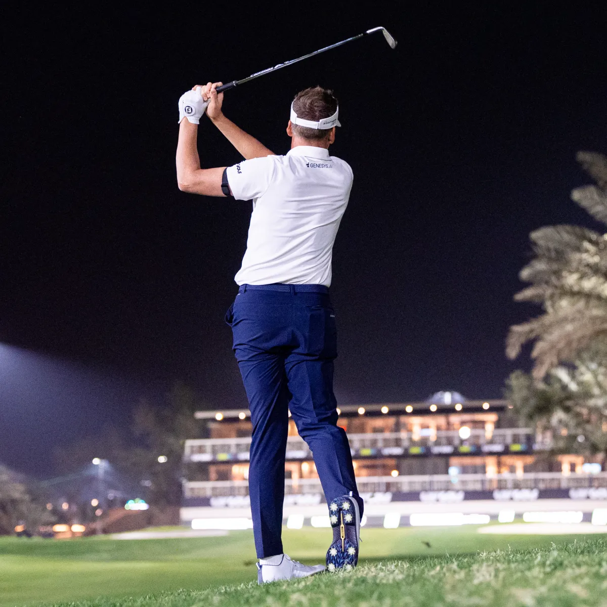 Golfer in a white shirt and blue pants finishes a swing on a night-lit course with palm trees and lit buildings in the background.