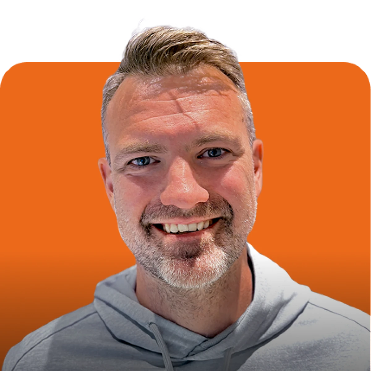 Close-up of a smiling man with short light brown hair and stubble, wearing a gray hoodie against an orange background.