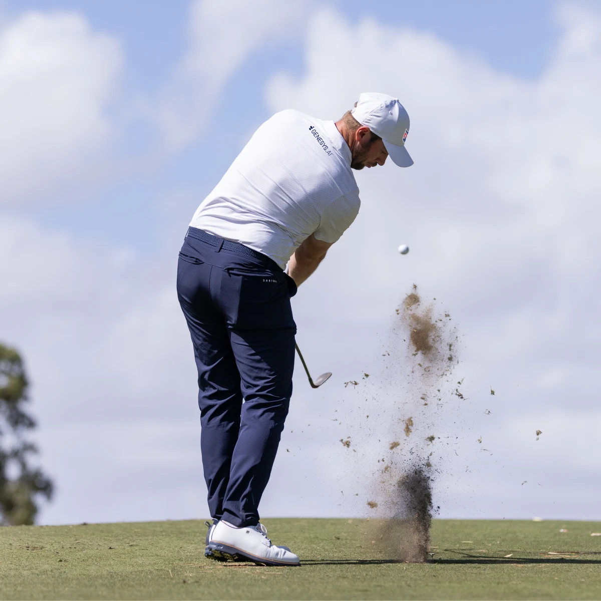 Golfer in a white shirt and cap, swinging a club on a grass course, with dirt flying and a blue sky background.