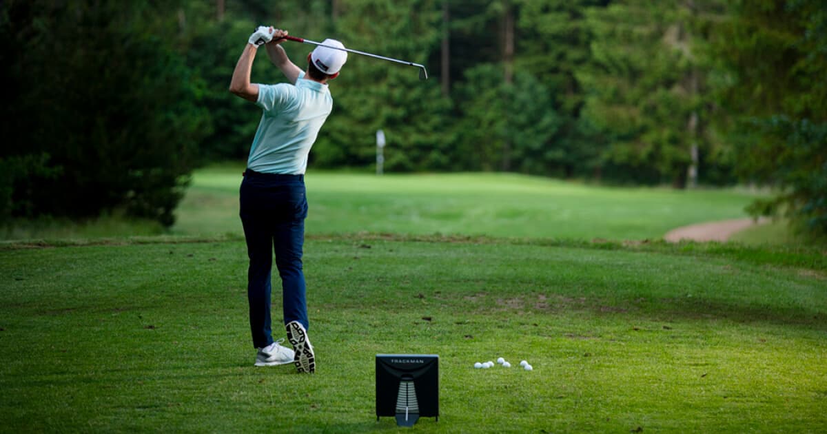 Golfer in a white cap and light blue shirt swings a club on a lush green course, with trees in the background and a device on the ground.