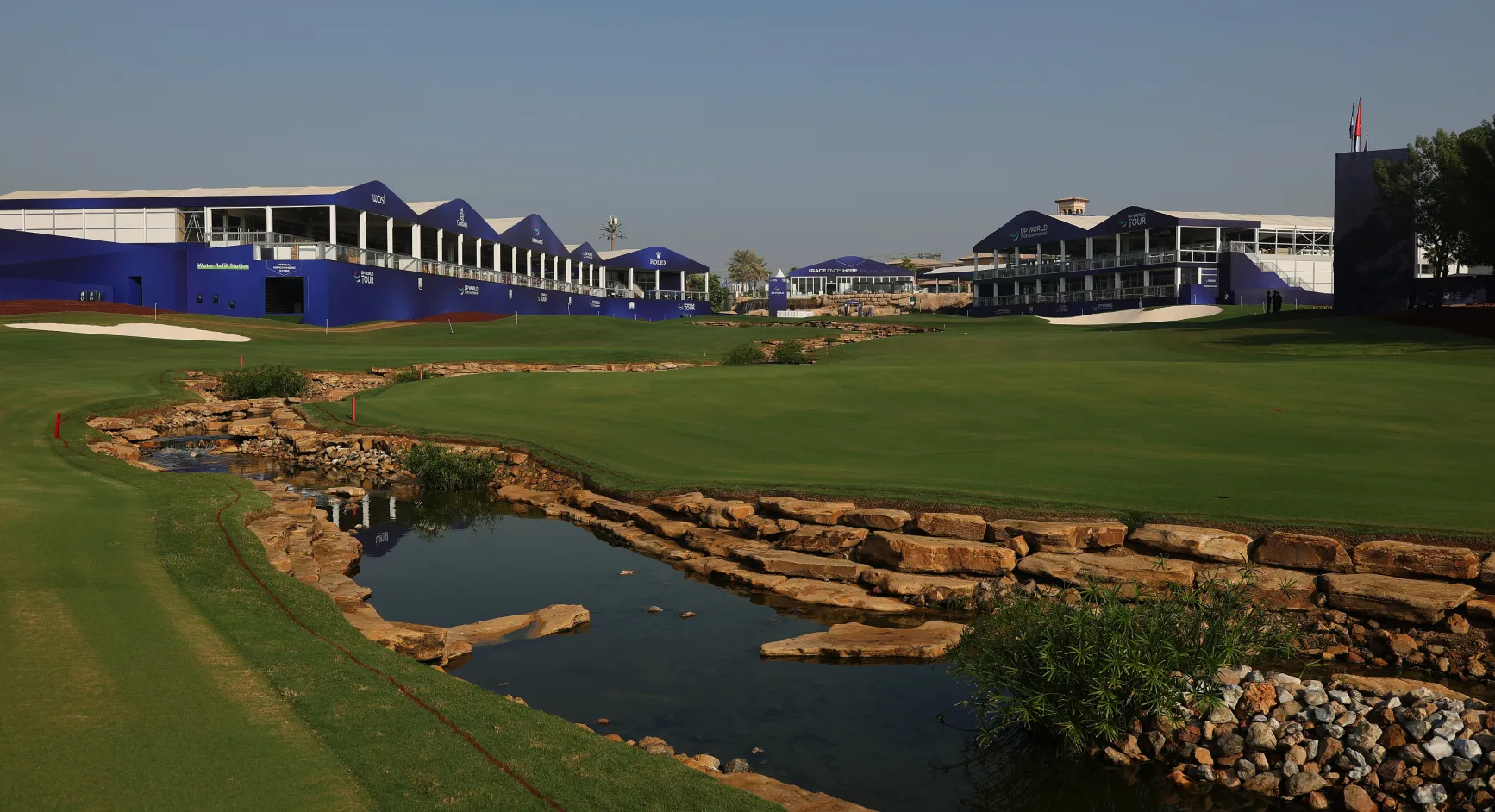 Golf course with lush green fairway, small pond with rocks, and large blue buildings in the background under a clear sky.