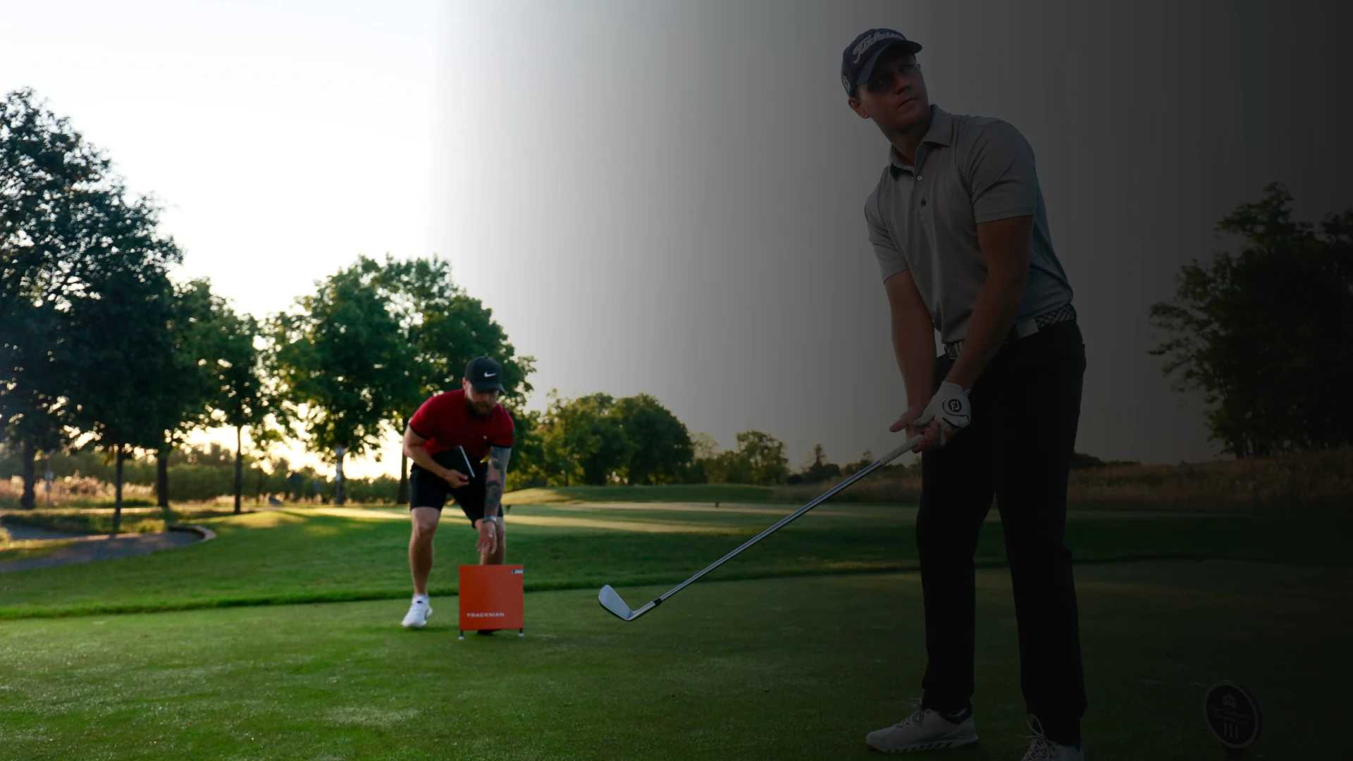 Two golfers on a course; one preparing to swing, the other adjusting a tee. Bright sun and trees in the background.