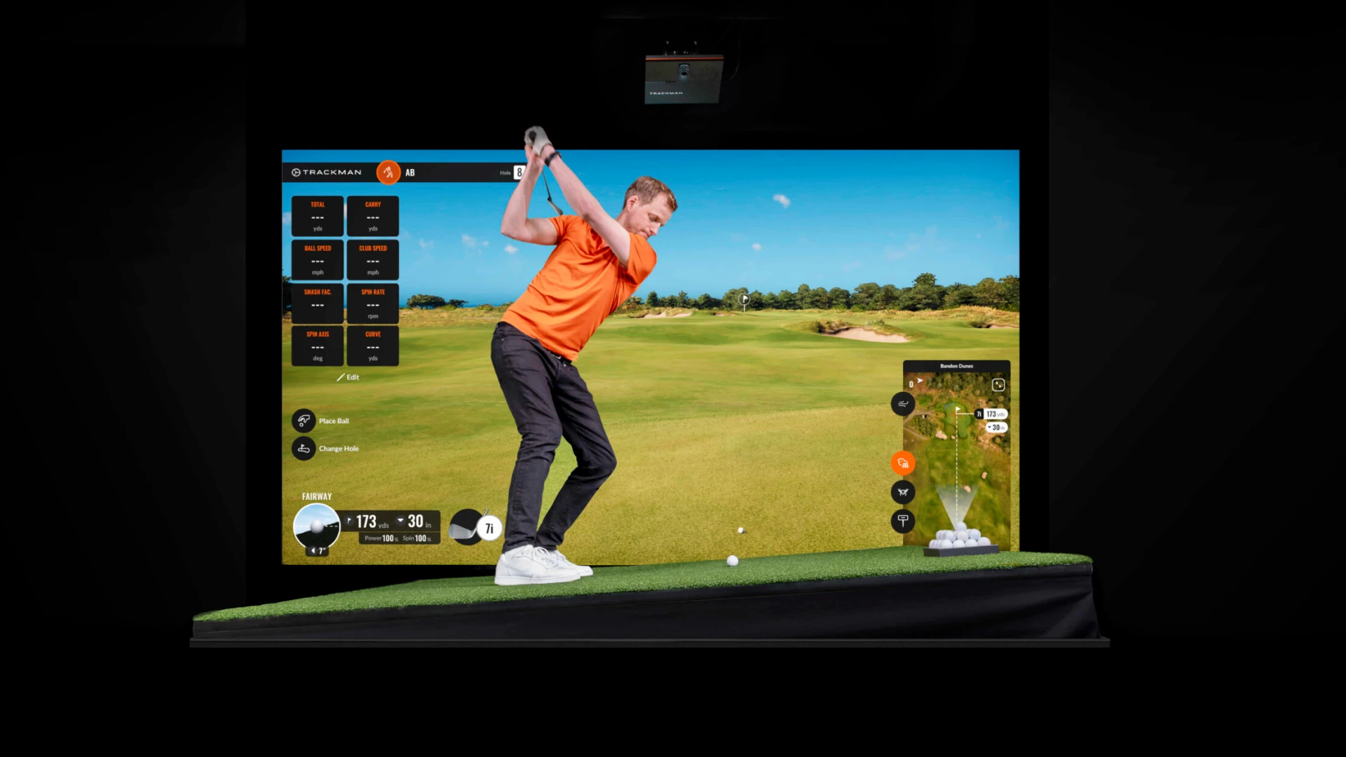 Golfer in orange shirt mid-swing on an indoor virtual golf simulator, showing a fairway scene with performance stats displayed.