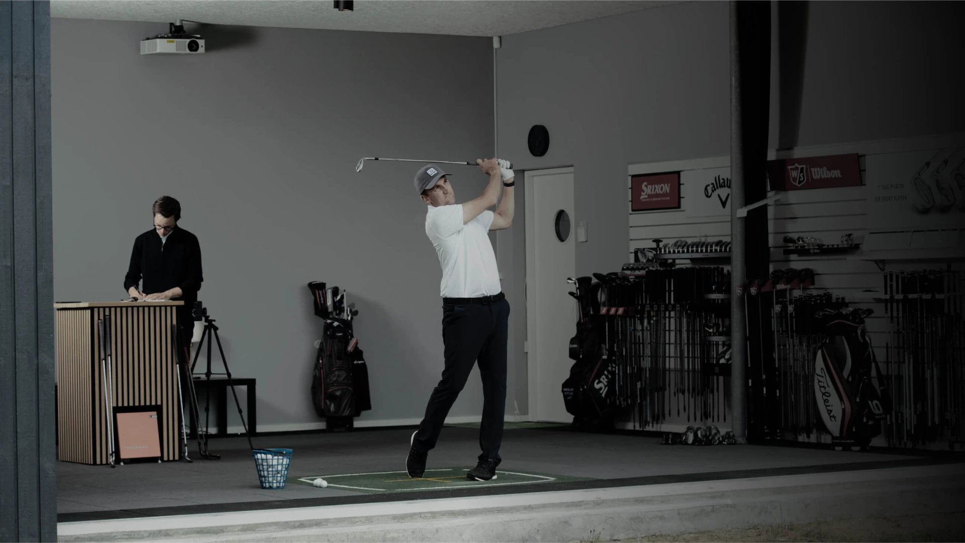 A golfer swings a club indoors, with a person observing data on a laptop. Golf bags and equipment are visible in the background.