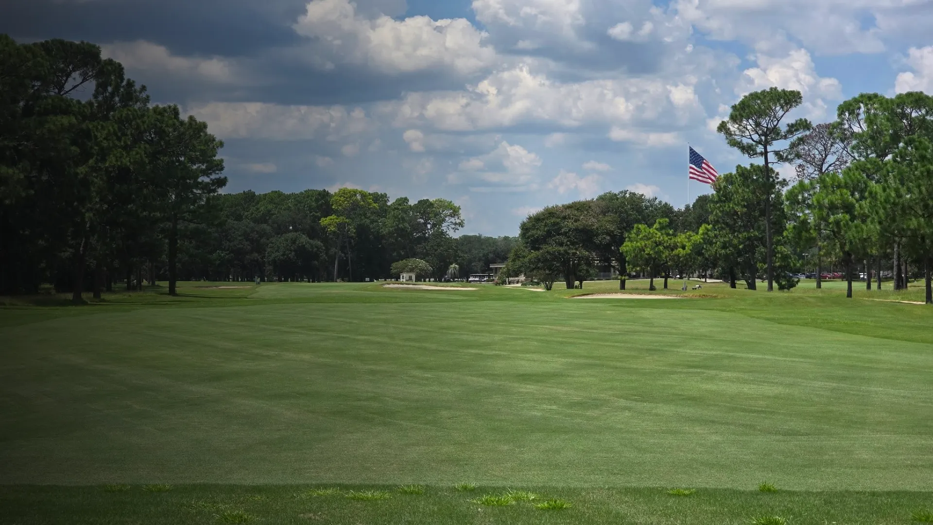 A well-maintained golf course with a large American flag waving between trees under a partly cloudy sky.
