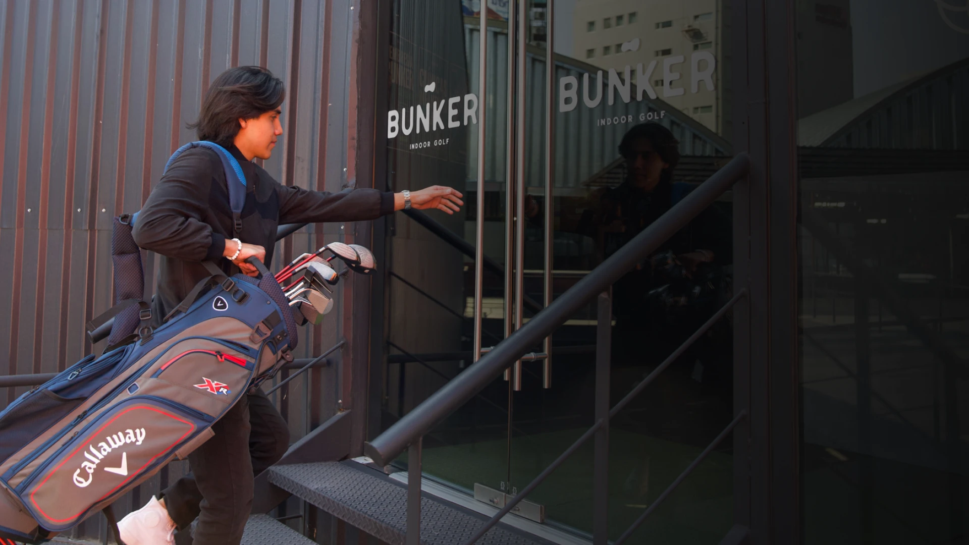Person carrying a golf bag enters a building labeled "Bunker Indoor Golf," with a modern exterior and reflective glass door.