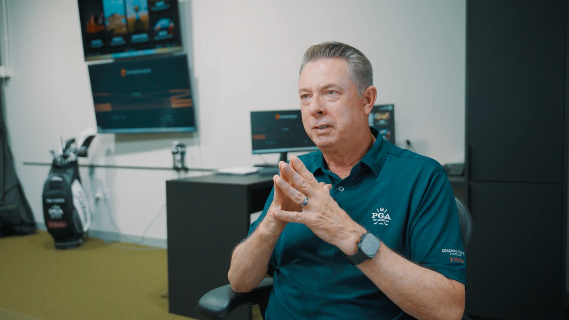 Man in a PGA shirt sits in an office with computer monitors and golf equipment in the background, speaking thoughtfully.