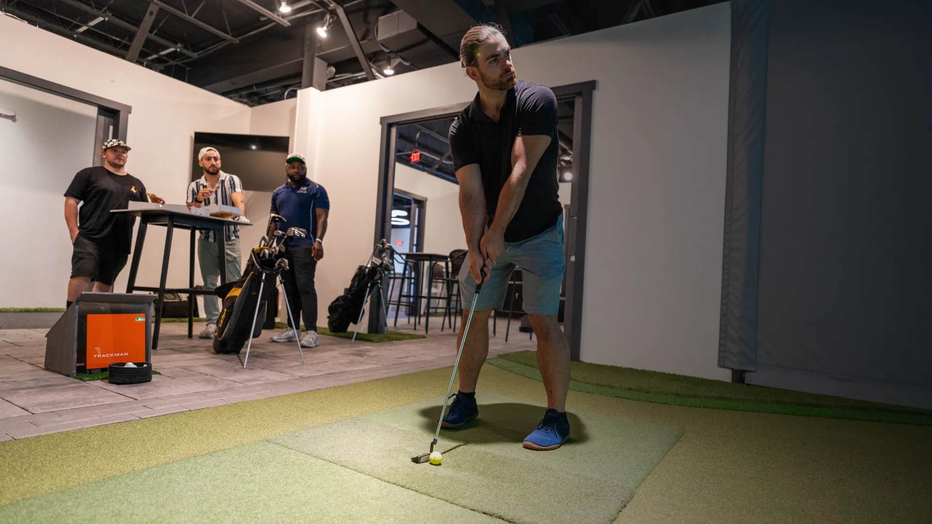 A man prepares to swing a golf club indoors while three others watch near a display screen and golf bags.