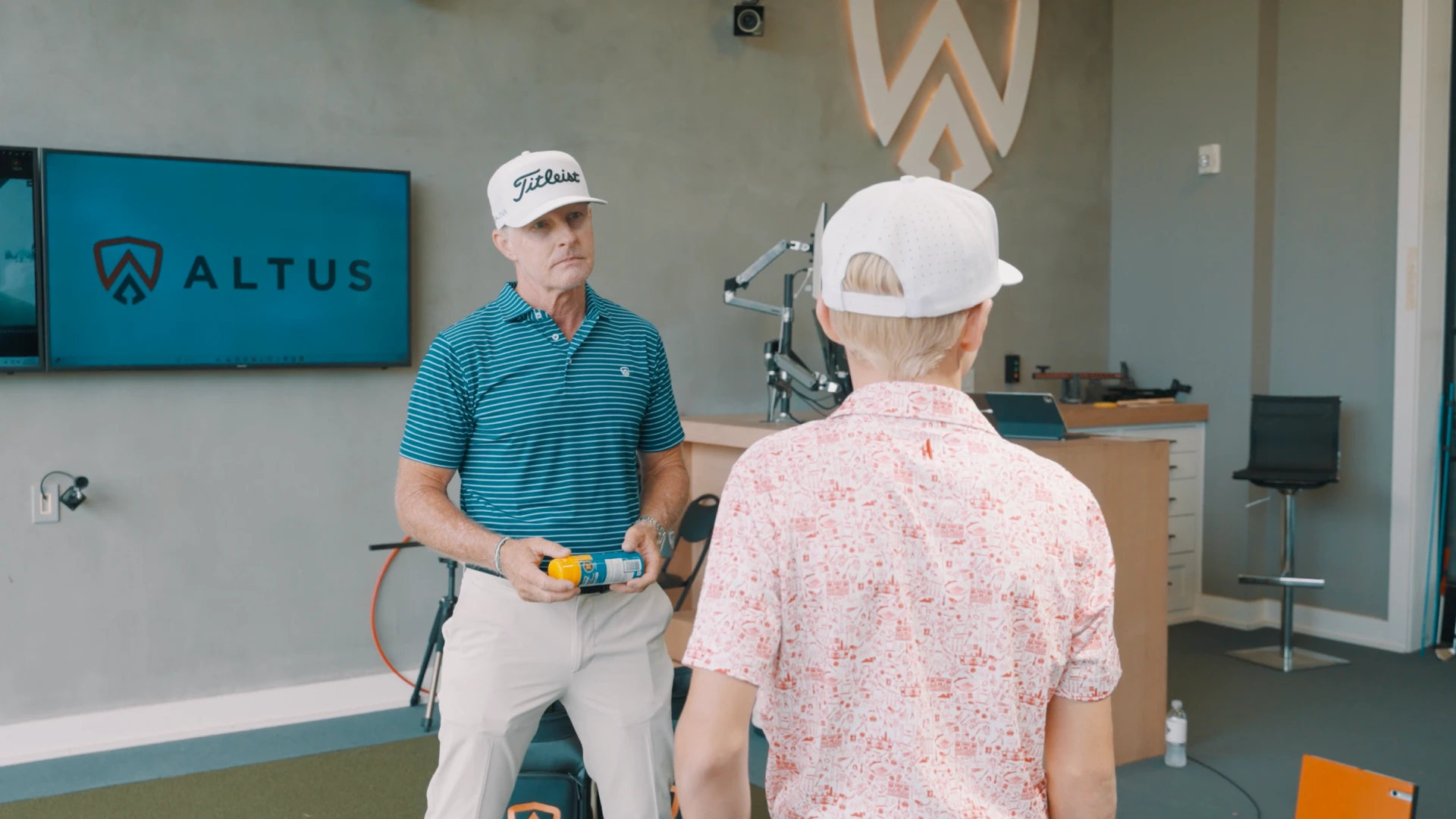 Two people in a golf training room, one holding a spray can, with an Altus sign and golf equipment in the background.