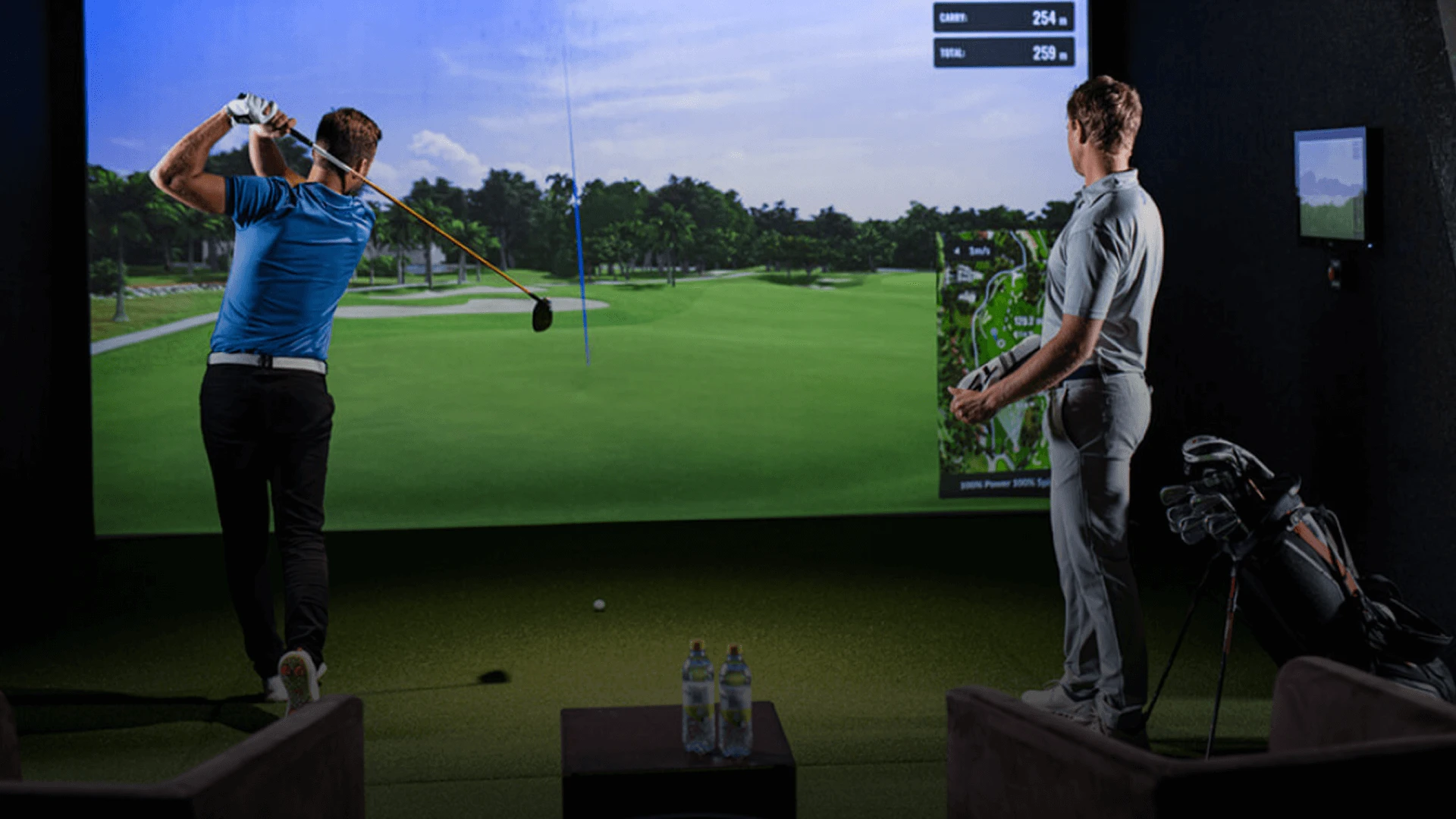 Two men playing indoor golf. One swings a club, while the other watches. A golf simulator screen displays a course. Golf bags and bottles nearby.