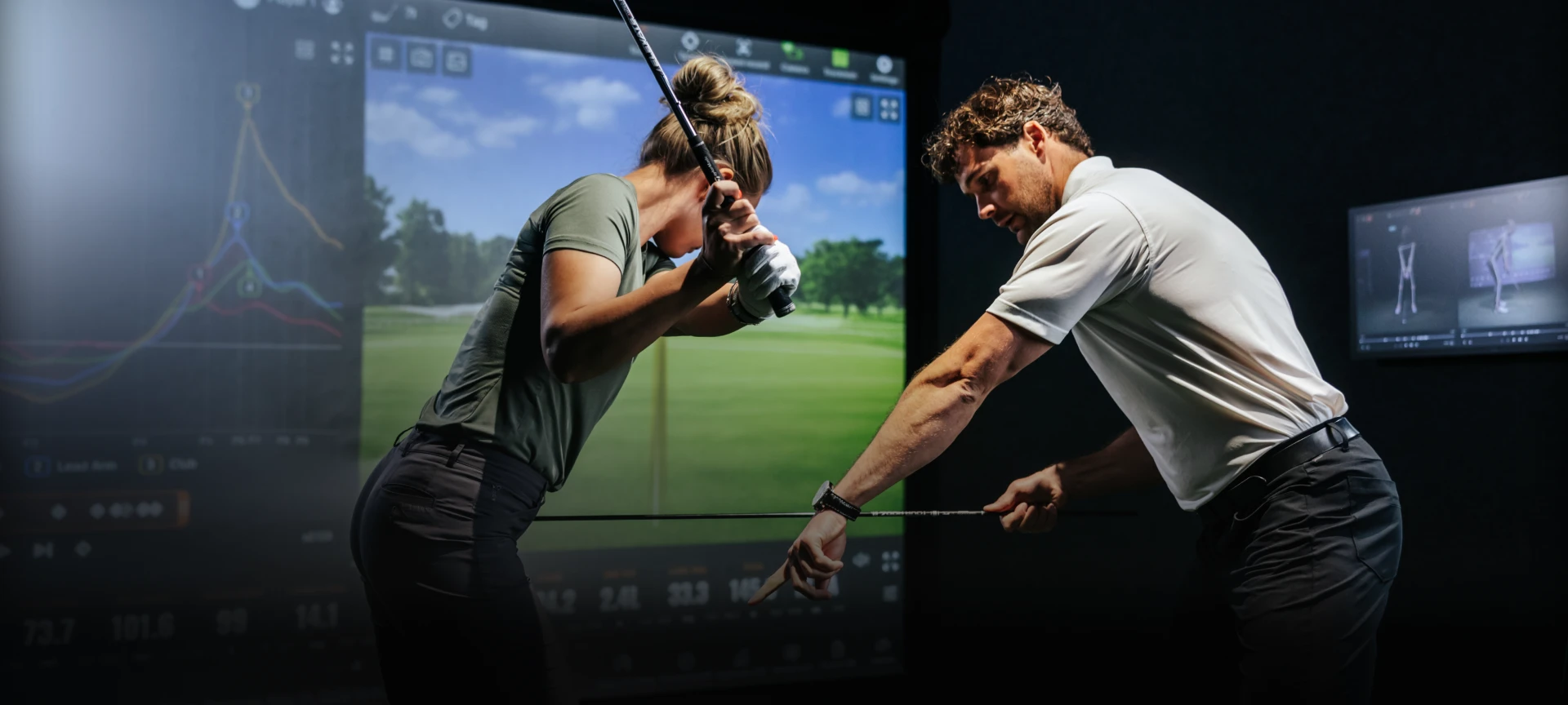 A golf instructor demonstrates a swing to a student in a golf simulator room with digital analysis displayed on screens.