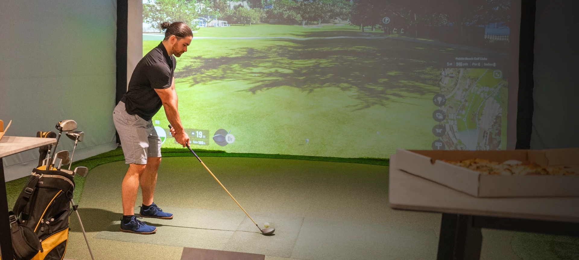 A man playing golf indoors using a simulator screen, with a golf bag and clubs nearby.