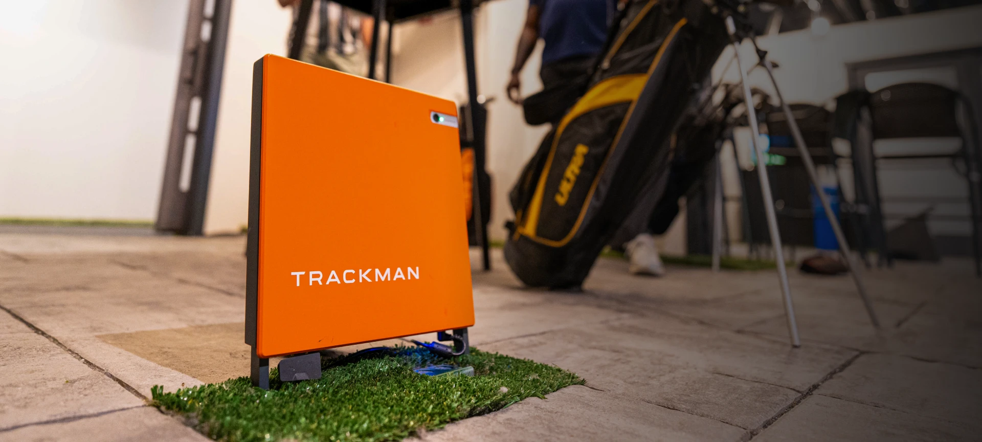 An orange Trackman device on artificial turf, with a blurred background showing a person and a golf bag indoors.