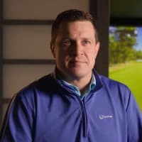 Man in a blue jacket standing indoors with a blurred background showing greenery and a bright sky.