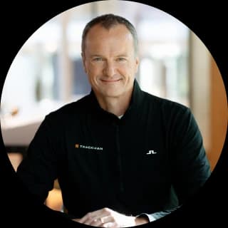 Smiling man in a black jacket sits indoors, natural light in the background.