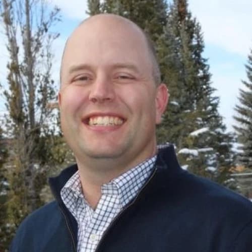Smiling man wearing a checkered shirt and dark jacket, standing outdoors with evergreen trees in the background.