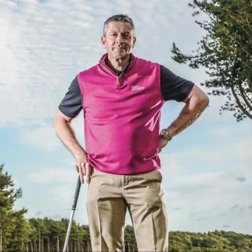 Man standing on a golf course, wearing a pink vest and holding a golf club, with trees and a cloudy sky in the background.