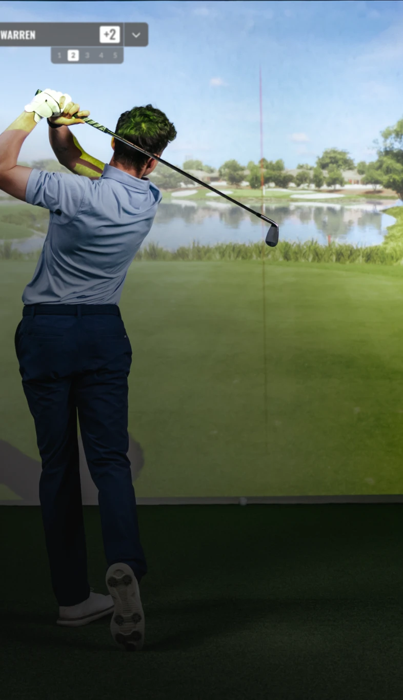 A golfer in mid-swing facing a virtual golf course, with a lake and trees in the background, wearing a blue shirt and dark pants.