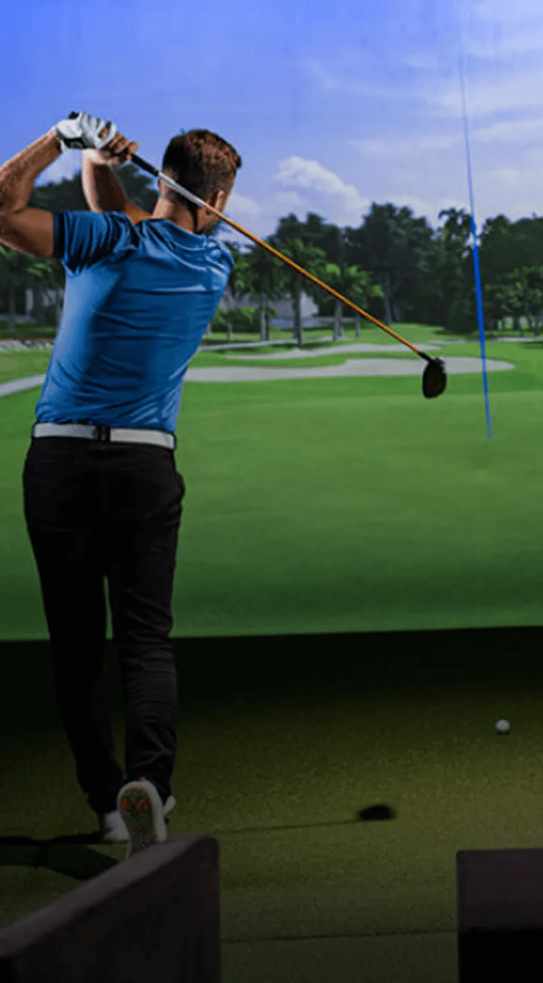 A golfer in a blue shirt swings a club in an indoor golf simulator, with a green course displayed on the screen in front of him.