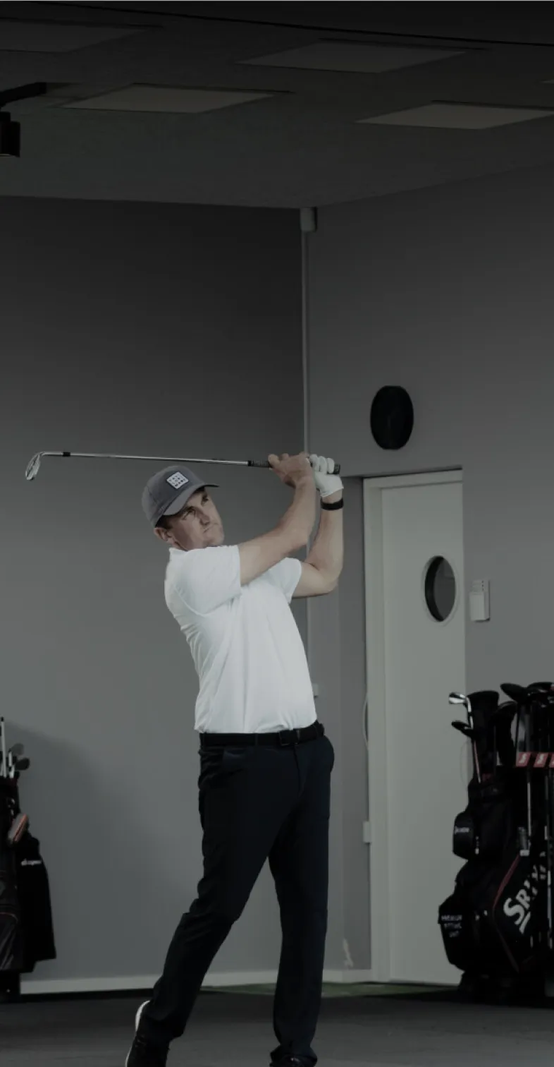 A person in a white shirt and cap swings a golf club indoors, surrounded by golf bags.