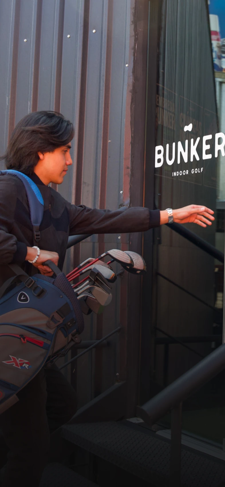 Person entering "Bunker Indoor Golf" while carrying a golf bag with clubs, reaching towards the door handle.