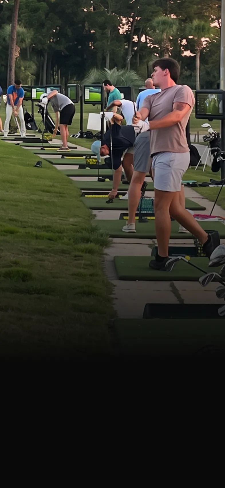 Golfers practicing swings at a driving range, with several people lined up on mats holding clubs, surrounded by green grass.