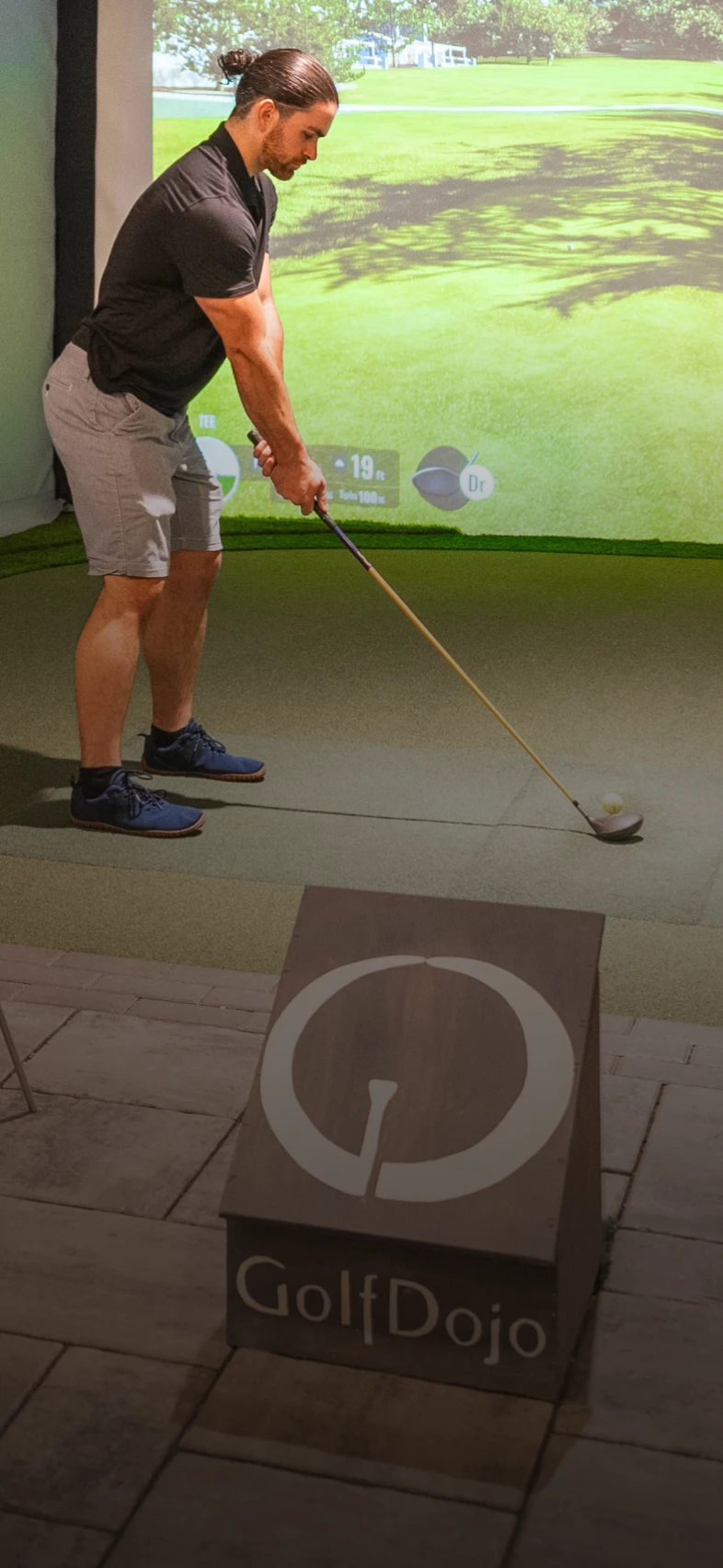 A man in casual attire stands on an indoor golf simulator, preparing to swing a golf club. A "GolfDojo" sign is visible in the foreground.