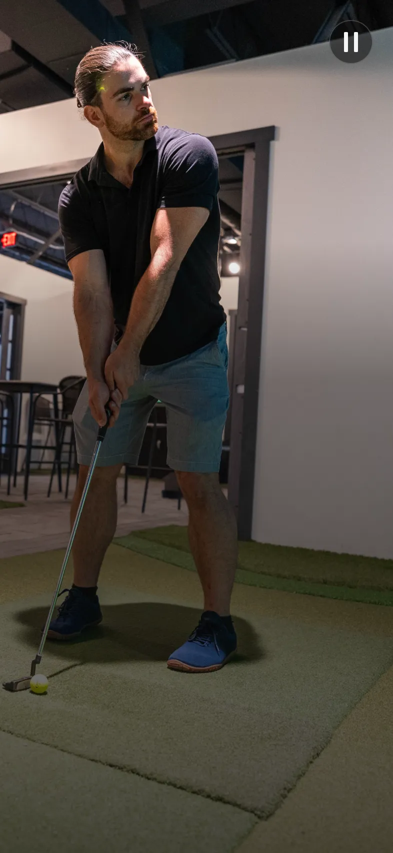 A man in a black shirt and gray shorts prepares to putt a yellow golf ball indoors on a green mat.