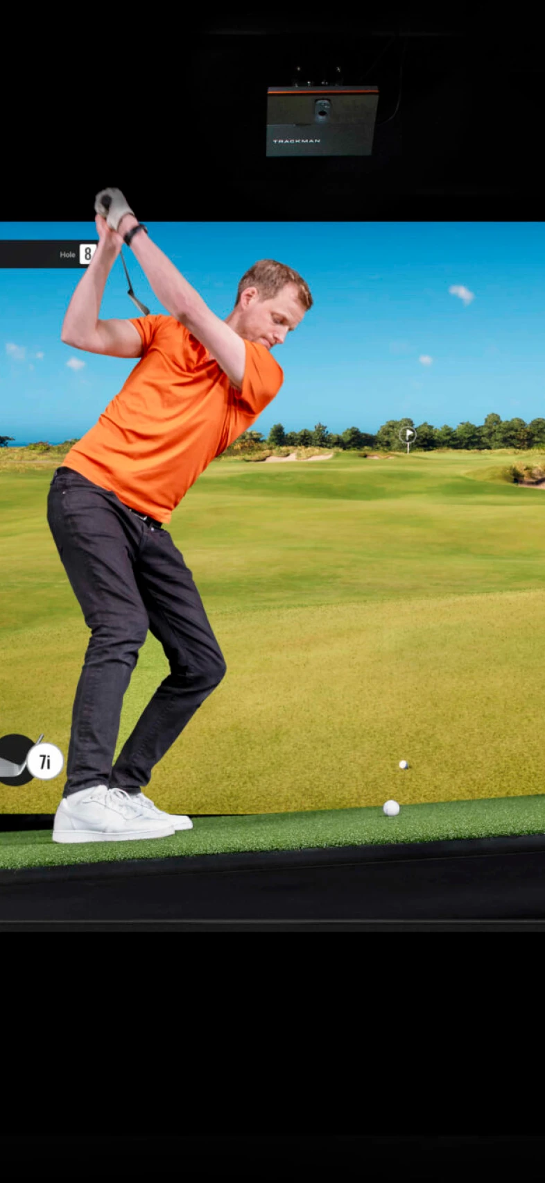 A golfer in an orange shirt prepares for a swing on a golf simulator, showing a virtual course backdrop.