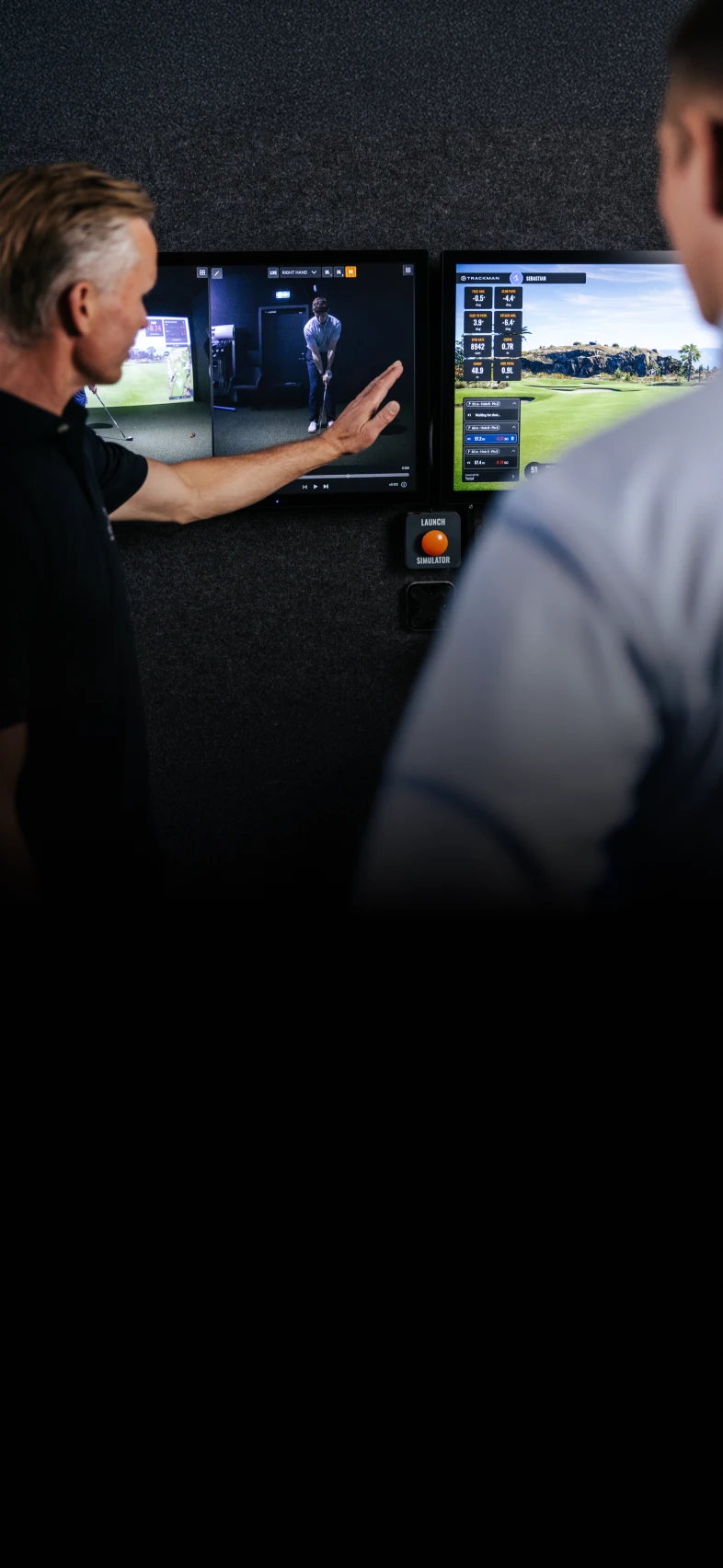 A man points at a video of a golfer on a screen while discussing with another person in front of golf performance data displayed on a monitor.