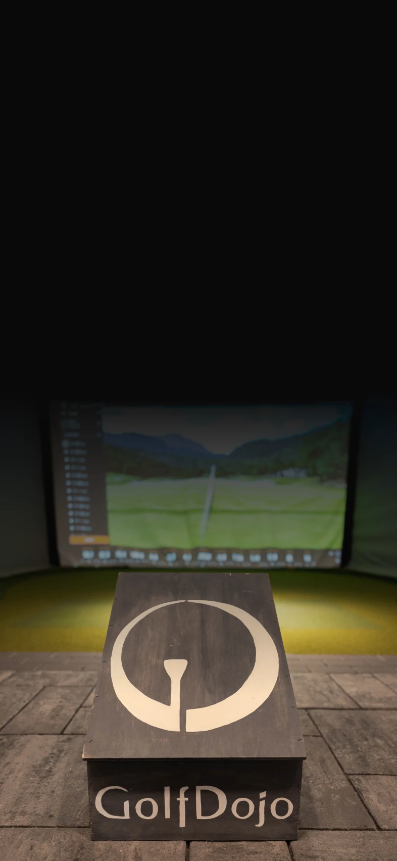 A golf simulator setup with a screen displaying a course scene and a wooden platform labeled "GolfDojo" in the foreground.