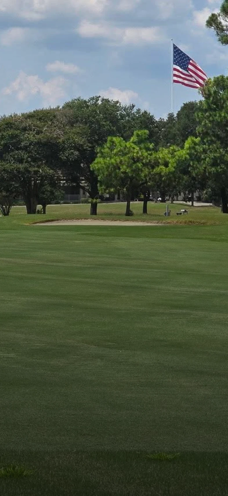 Lush green golf course with trees in the background under a partly cloudy sky. An American flag waves from a tall pole amid the trees.