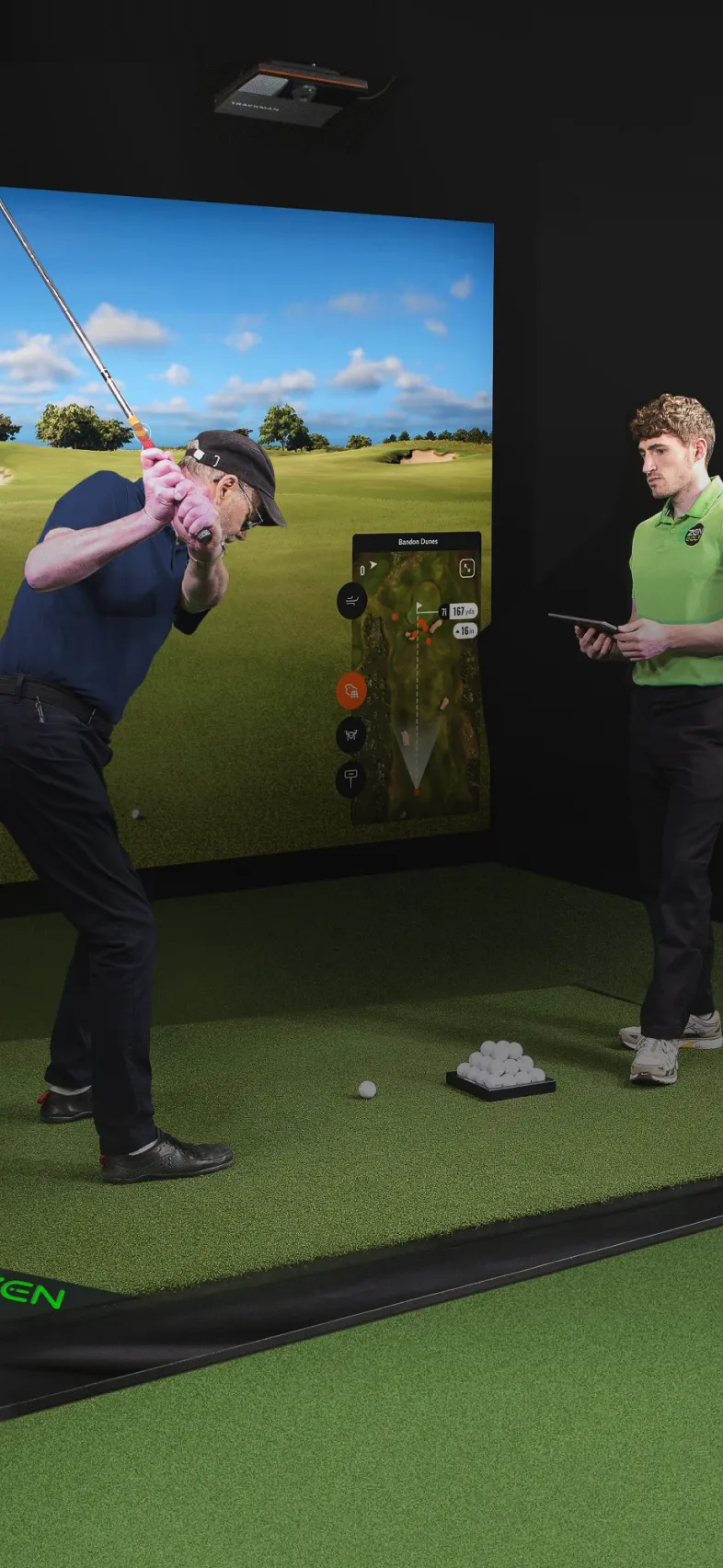 Golfer practicing swing indoors with a simulator screen. An instructor stands nearby, holding a tablet, observing the swing.