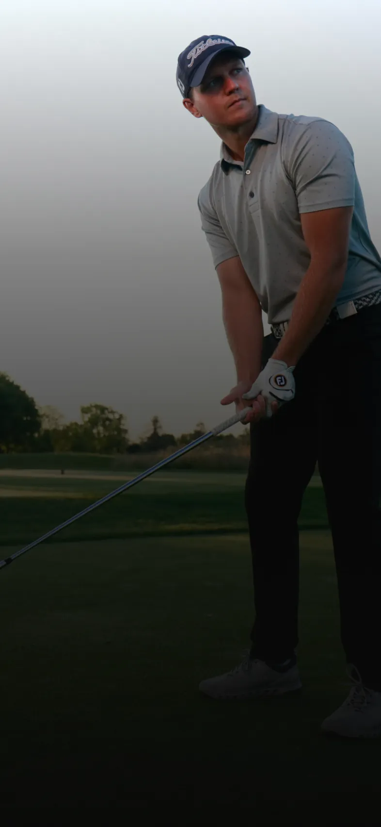 Man in a gray polo and cap prepares to swing a golf club on a course at dusk. Trees and a clear sky are in the background.