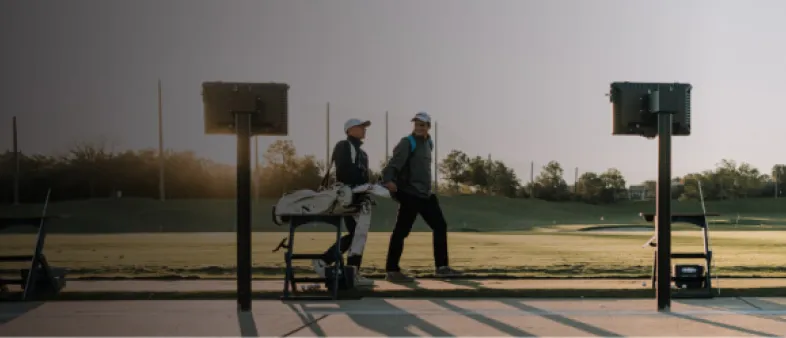 Two golfers walk on a driving range at sunset, carrying clubs. Golf bags rest on stands in the foreground.