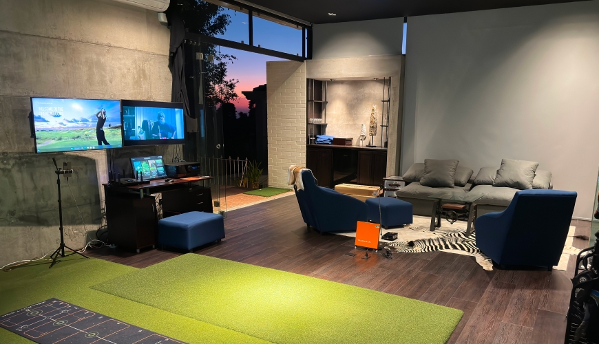 A trackman home indoor golf simulator, two screens, green mat, blue chairs, sofa, and sunset view through large windows. Cozy and tech-savvy ambiance.